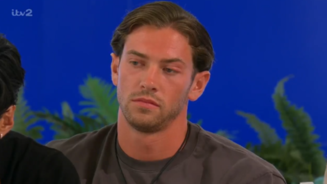 A man with brown hair and a beard, wearing a dark grey t-shirt, looks slightly to the left with a serious expression.
