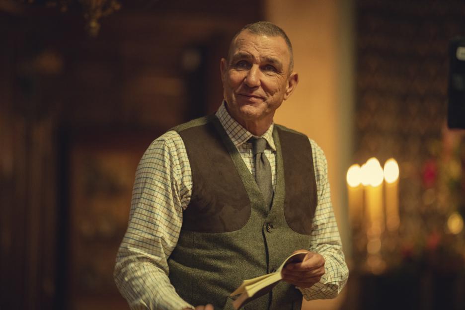 A man in a plaid shirt and vest smiles while holding a small book.