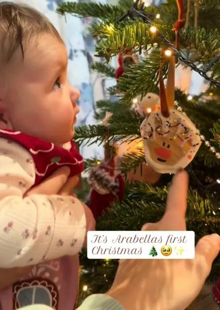 A baby girl held by a parent looking at a homemade salt dough ornament shaped like a reindeer on a Christmas tree.