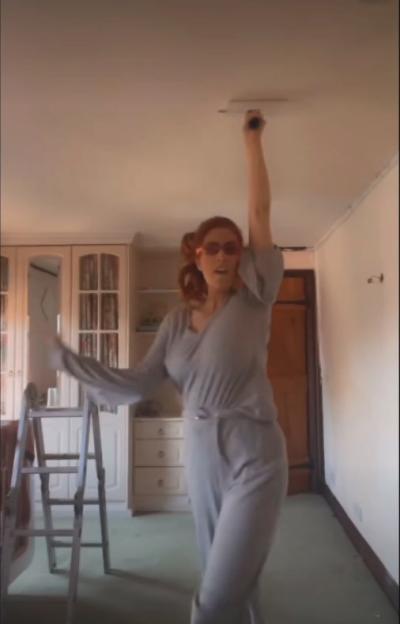 A woman in a gray jumpsuit and sunglasses reaching overhead to clean a ceiling light fixture.