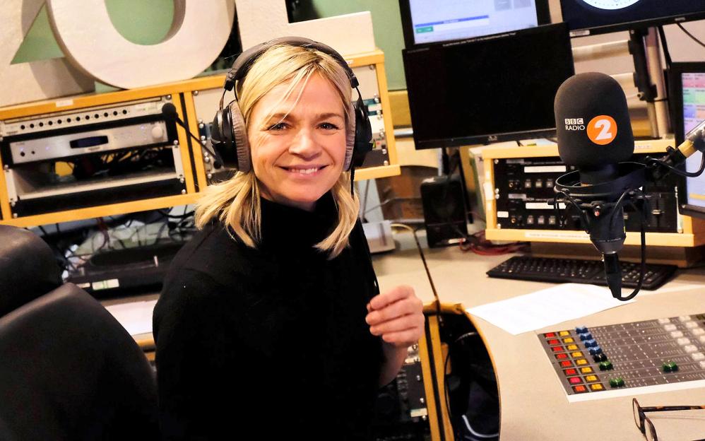 Zoe Ball, a BBC Radio 2 DJ, smiles while wearing headphones at a mixing desk with a BBC Radio 2 microphone.