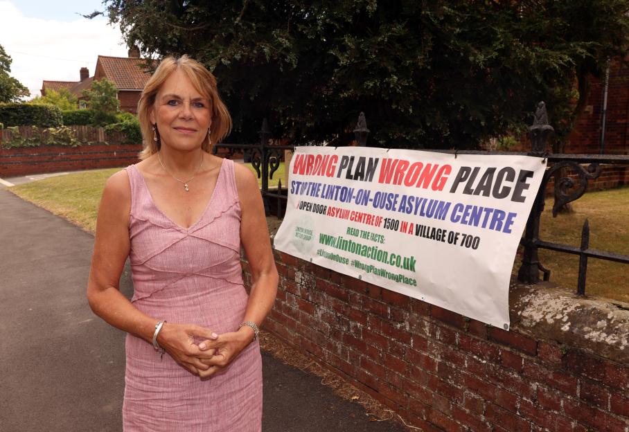 Professor Olga Matthias standing next to a protest banner that reads "WRONG PLAN WRONG PLACE, STOP THE LINTON-ON-OUSE ASYLUM CENTRE, OPEN DOOR ASYLUM CENTRE OF 1500 IN A VILLAGE OF 700".