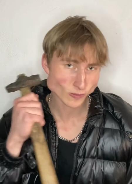A young man with light brown hair and a silver chain necklace wears a black puffer jacket and holds a hammer, looking at the camera with an intense expression.
