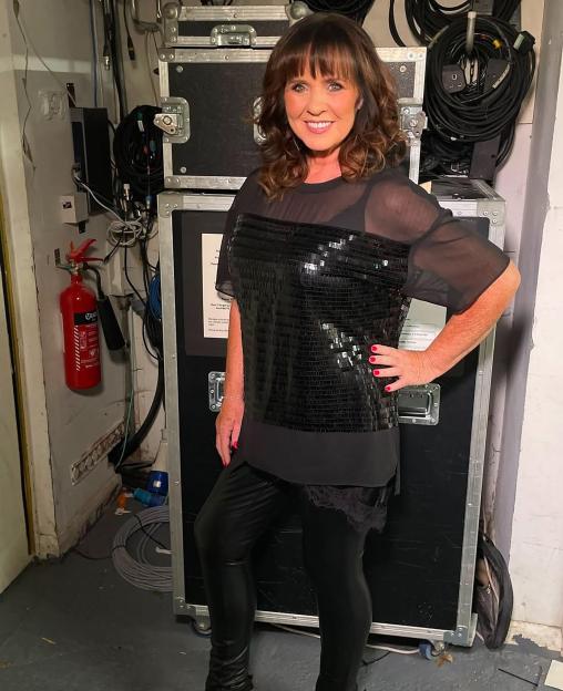 Coleen Nolan smiling, wearing a black sequined top and leather pants, posing backstage.
