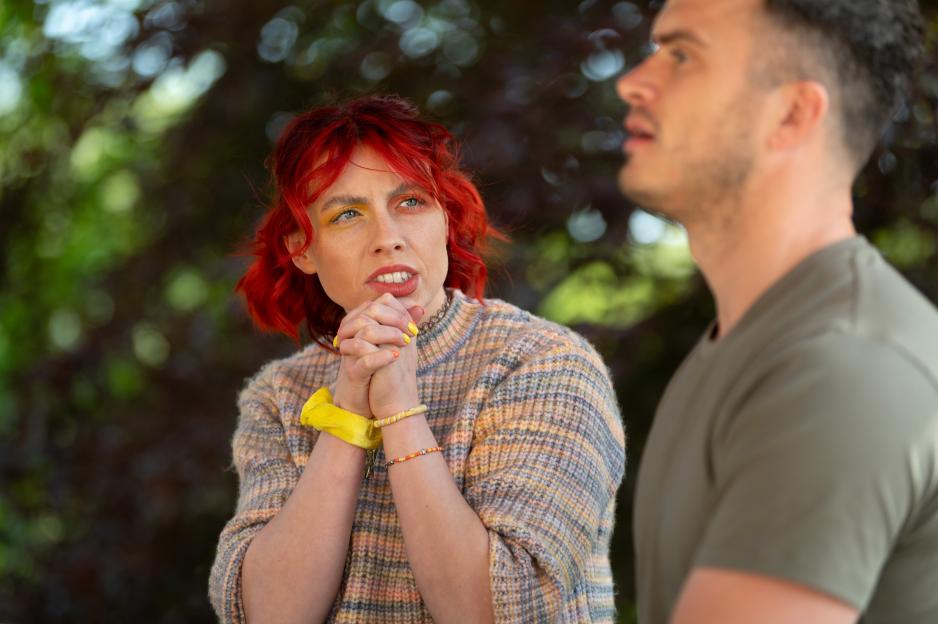 Jessie, a contestant on the BBC show "The Traitors," with hands clasped as if in prayer, looks worriedly toward fellow contestant Matthew, out of focus to the right.