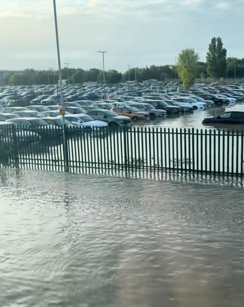 Video still of a flooded car park at Heathrow Airport, with numerous cars partially submerged in water behind a metal fence.
