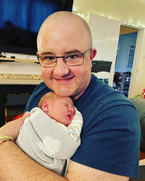 A man with glasses smiling while holding a swaddled newborn baby.