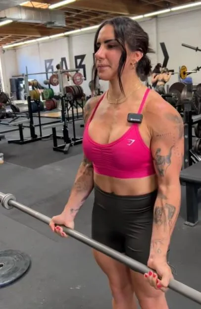A muscular woman wearing a pink sports bra and black shorts holds a barbell in a gym.
