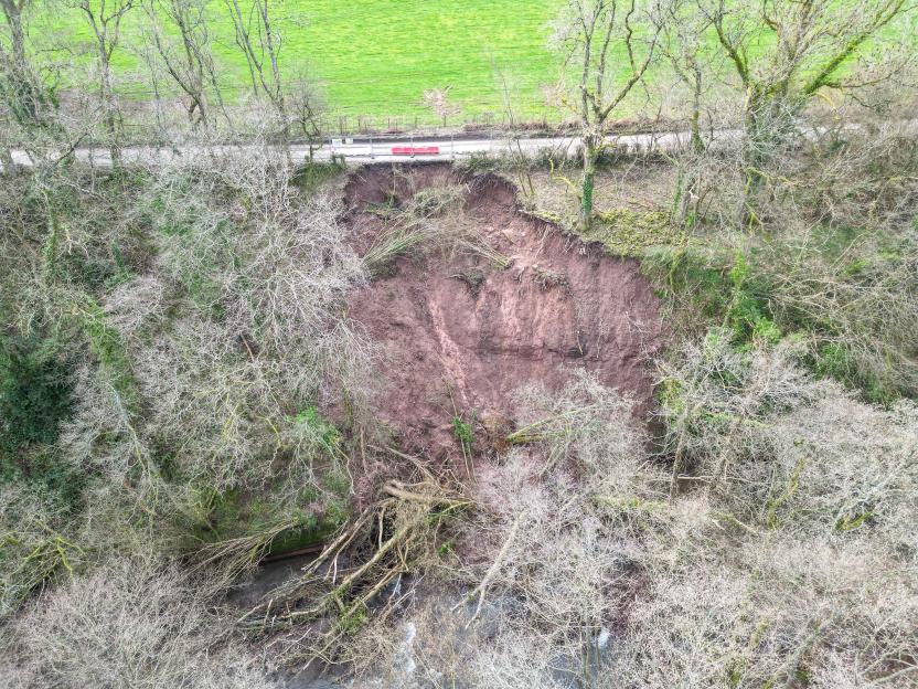 An aerial view of a massive landslip that has caused a road to collapse into a ravine, leaving a large exposed dirt cliff face and trees toppled into a river below.