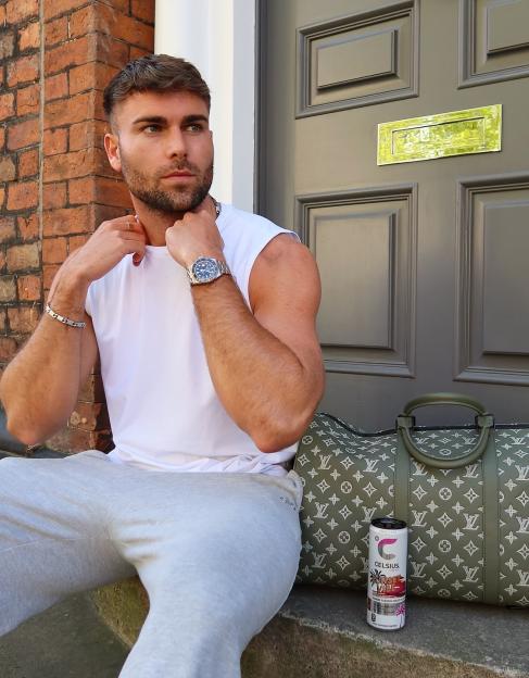 Man in a white tank top and grey sweatpants sitting on a doorstep with a Louis Vuitton duffle bag and a Celsius energy drink.