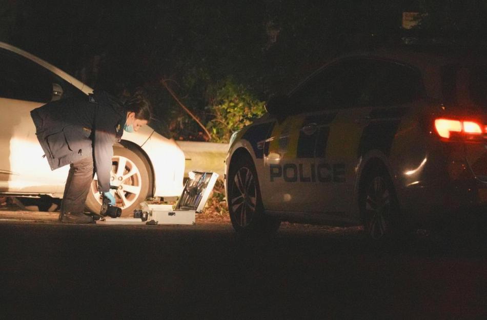 A forensic officer at night, illuminated by a car's headlights, inspects a crime scene next to a police car.