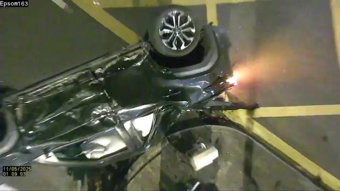 Overhead view of a crashed dark gray Toyota Yaris, flipped onto its side, with sparks coming from the crumpled front.