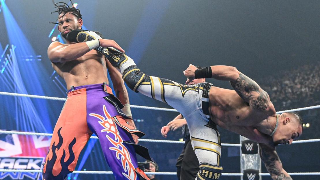A wrestler in purple and orange pants holds another wrestler in white and gold gear by the neck.