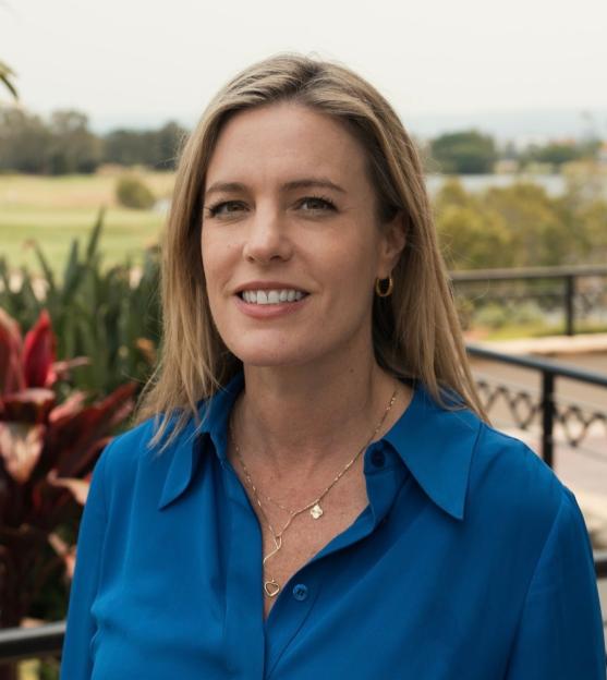 A smiling blonde woman in a blue shirt with a gold necklace and hoop earrings, standing outdoors.