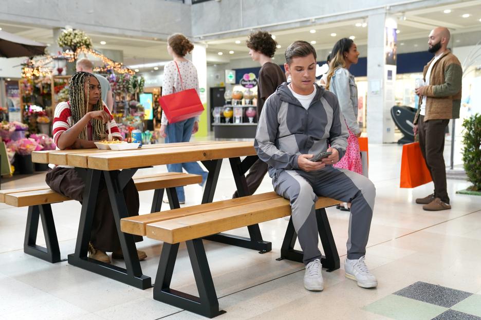 Two people sitting at a picnic table in a shopping mall with other shoppers in the background.
