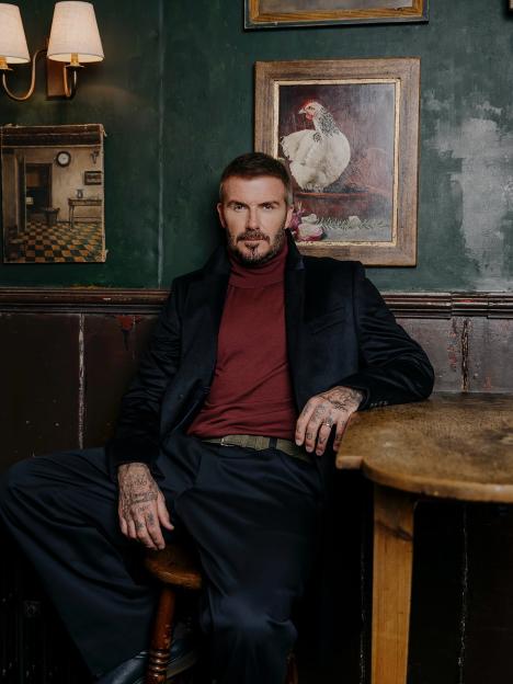 David Beckham wearing a red turtleneck, a black coat, and black pants, sitting on a stool at a table.