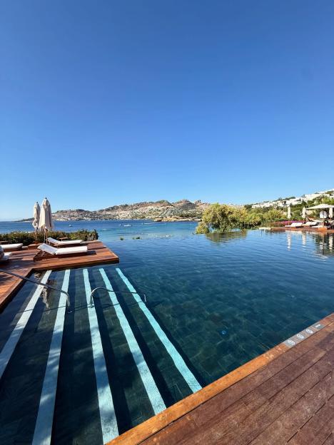 Infinity pool overlooking a coastline with buildings and hills under a clear blue sky.