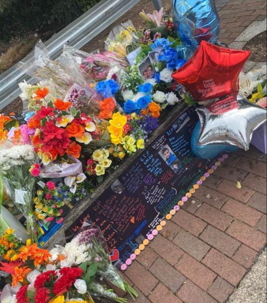 A roadside memorial with bouquets of flowers, balloons, and written tributes for a deceased son.