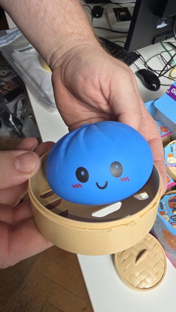 A person holding a blue squishy dumpling toy with a happy face in a plastic steamer basket.
