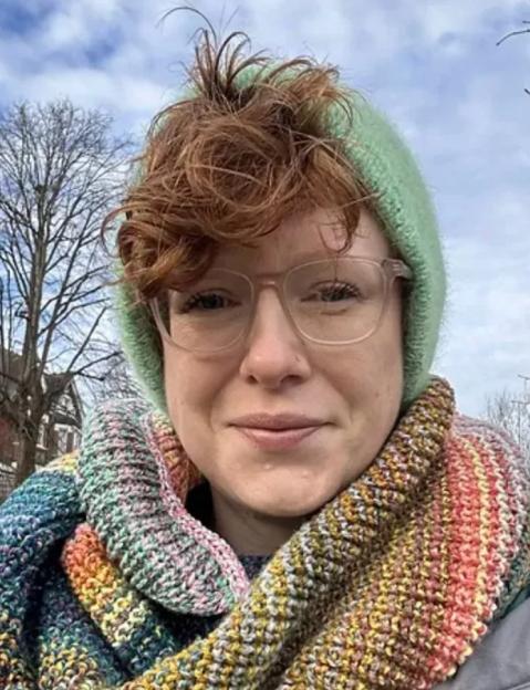A woman with reddish-brown hair, wearing glasses, a light green hat, and a colorful knitted scarf.