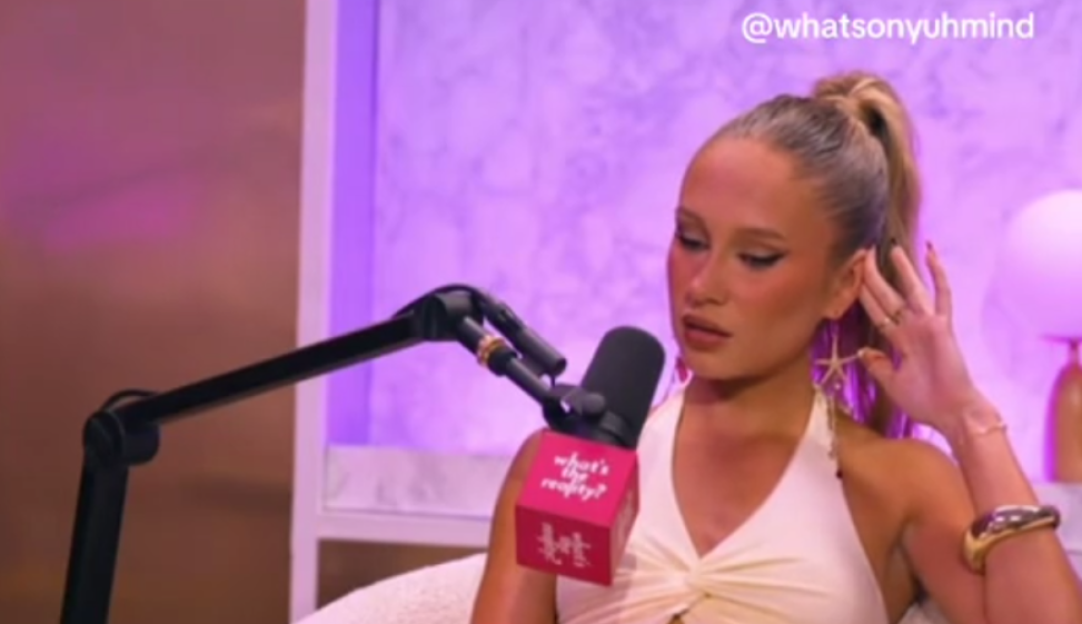 A woman with her hair in a high ponytail, wearing a white top and star-shaped earrings, speaks into a microphone with a pink foam cover that says "what's the reality?".