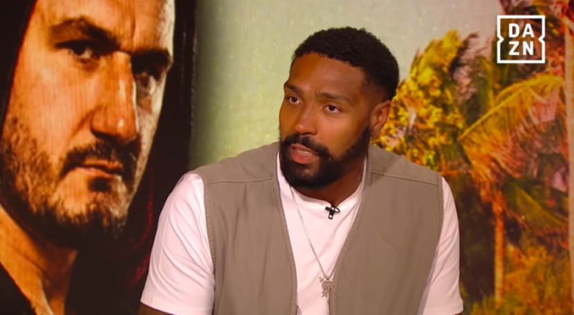 Man with beard and white t-shirt speaking on DAZN.