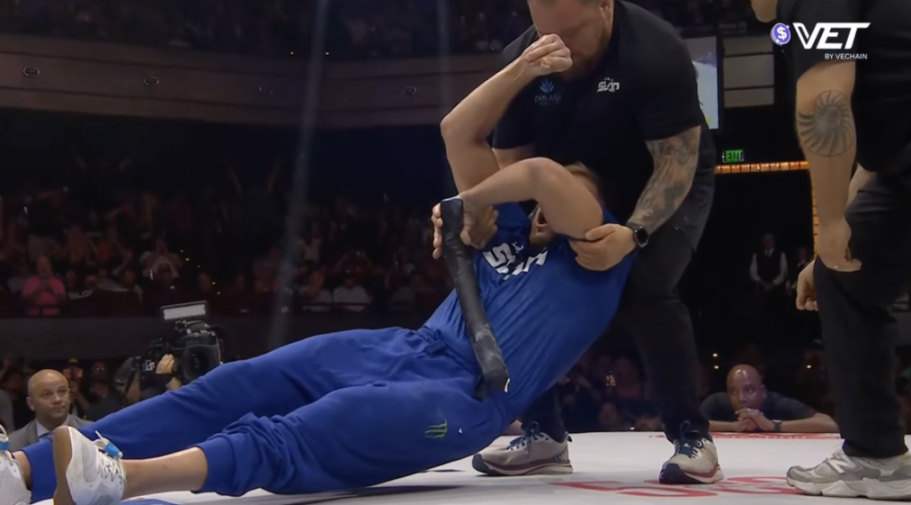 A man in a blue jumpsuit is held in a submission hold by another man while on the ground.