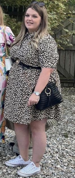 A person with long blonde hair, wearing sunglasses on her head, a leopard print dress, a black belt, a black shoulder bag, a silver watch, and white sneakers.