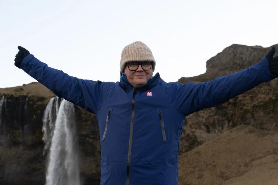 Alan Carr smiling and posing with arms outstretched in front of a waterfall.