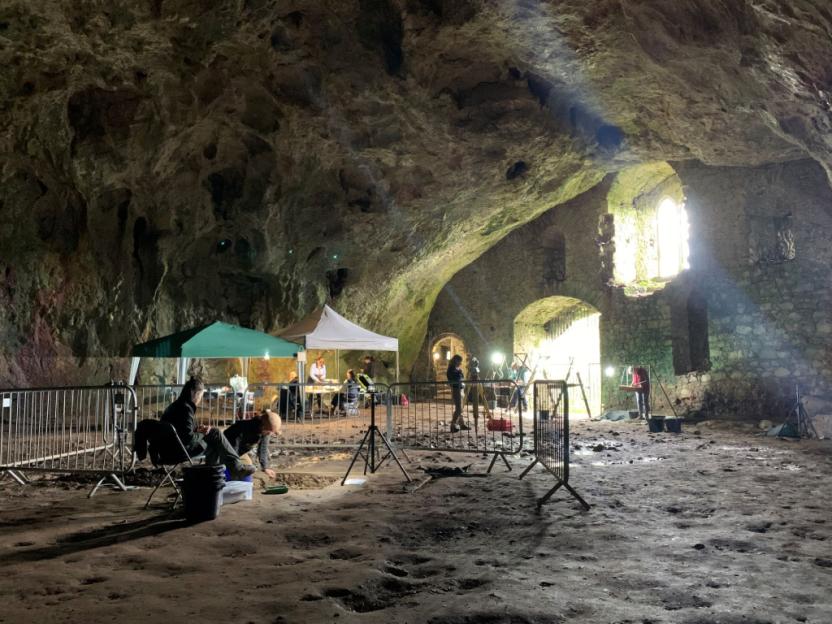 Archaeological excavation inside a cave, with several people working under tents and by stone walls.
