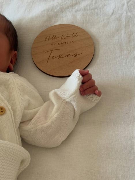 A baby in a white sweater lies next to a wooden disc engraved with "Hello World MY NAME IS Texas."