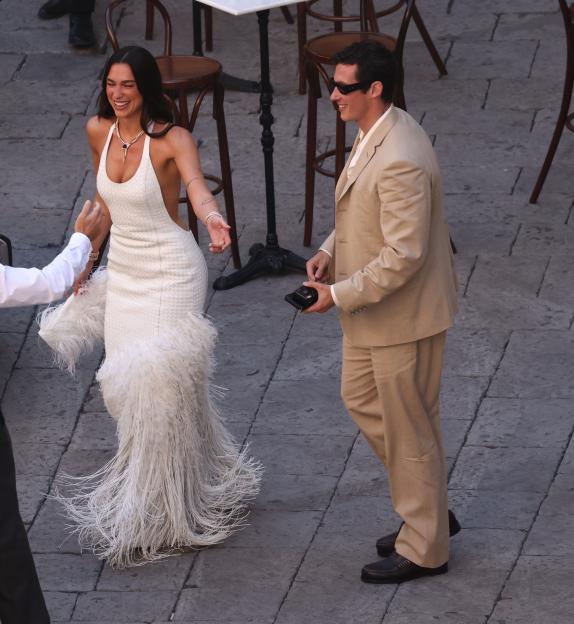 Dua Lipa in a white feathered dress smiles as she walks with Callum Turner in a tan suit and sunglasses.