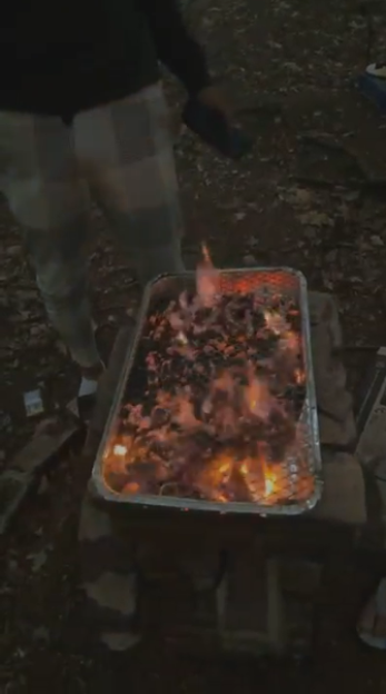 Foil pan filled with burning coals and embers, set on bricks on the ground.
