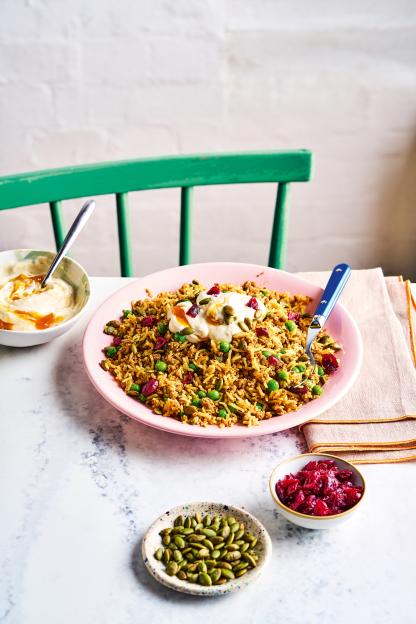 A turkey biryani dish with peas, pumpkin seeds, and cranberries, topped with yogurt and a drizzle of sauce, on a pink plate.