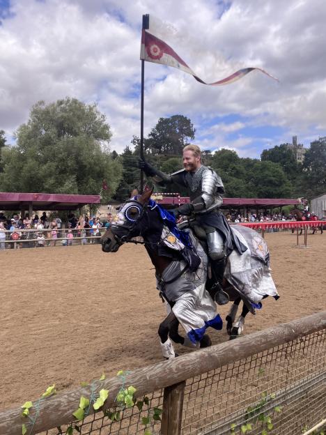 A smiling knight on horseback, dressed in shining armor and holding a banner, at a jousting tournament in Towcester.