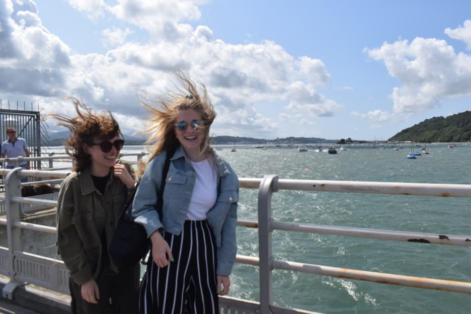 Two young women on a bridge over choppy water, with their hair blowing in the wind.