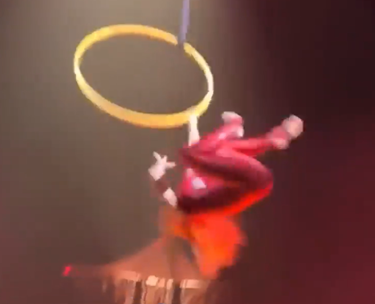 An acrobat in a red suit hangs upside down from a golden aerial hoop.