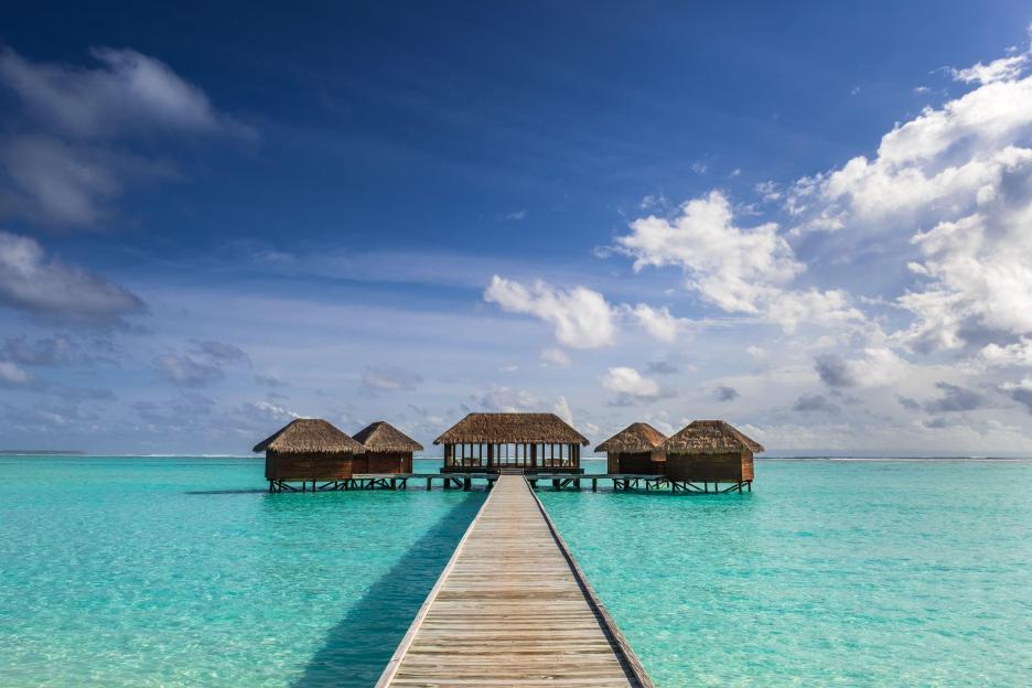A wooden boardwalk leading to several over-water bungalows with thatched roofs at a spa in the Maldives.