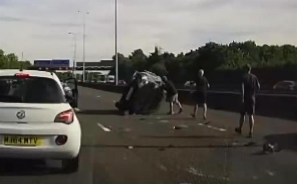 A crashed car overturned on a motorway with debris, while men stand nearby.