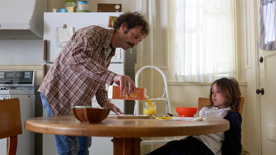 A man with a cigarette in his mouth pours orange juice for a young girl at a kitchen table.