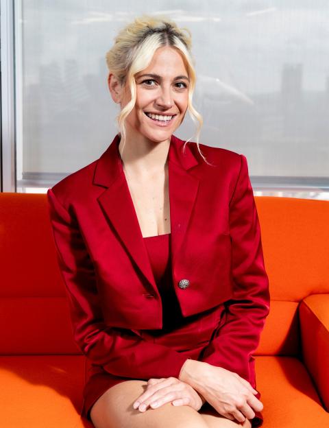Pixie Lott smiles while sitting on an orange couch, wearing a red suit.