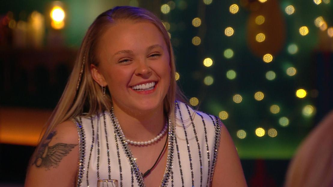 JoJo Siwa smiling while wearing a white top with silver sequins and a pearl necklace.