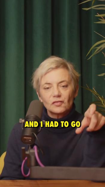 A woman with short, light-colored hair, wearing a dark blue top, speaks into a microphone while pointing with her right hand.