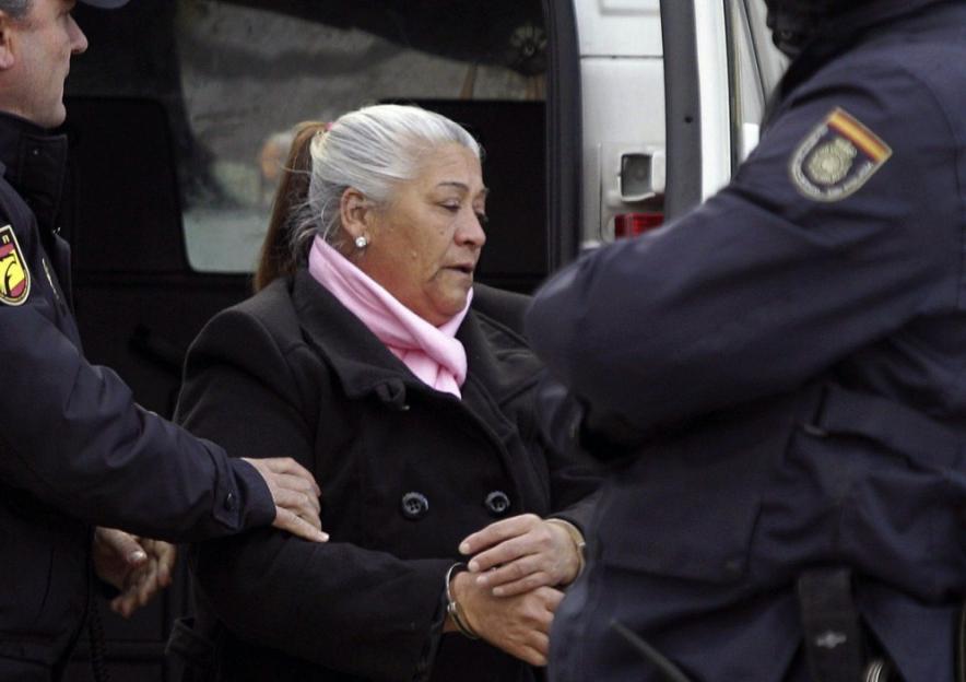 A handcuffed woman, wearing a black coat and pink scarf, is escorted by two police officers.