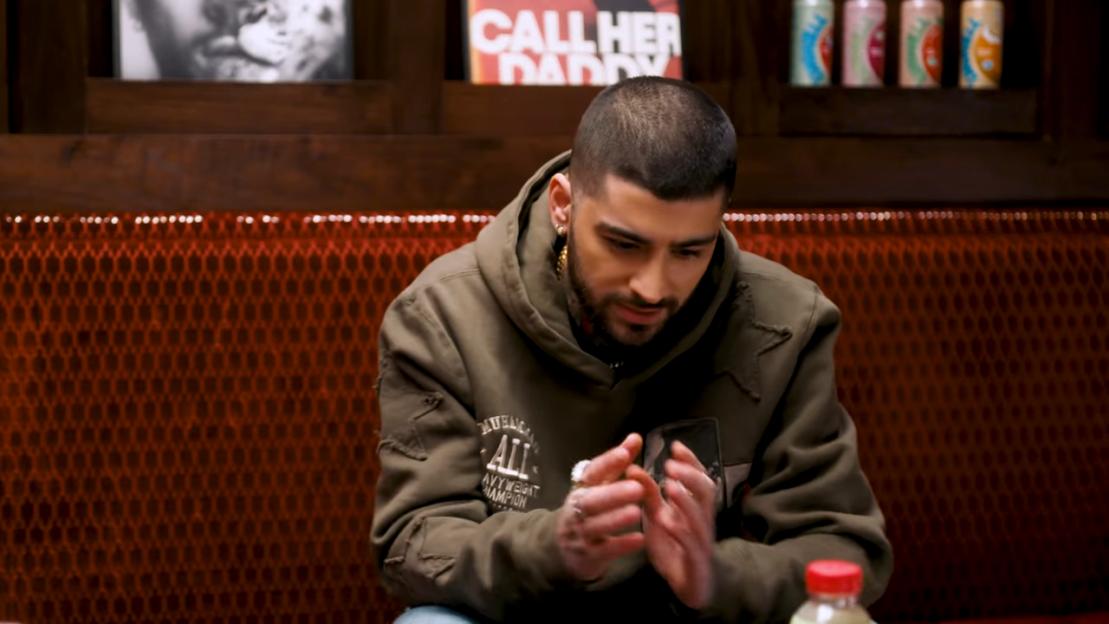 Zayn Malik looking down with his hands clasped, wearing a khaki hoodie, against a dark red patterned background.