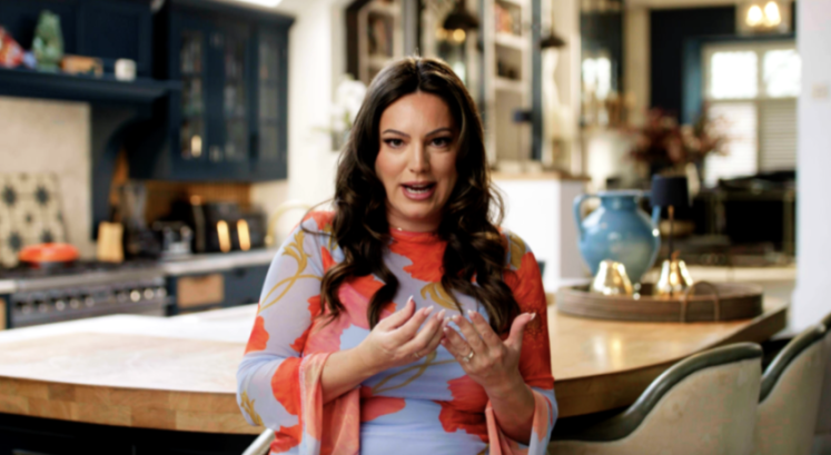 A woman in a floral mesh top speaking in a kitchen.