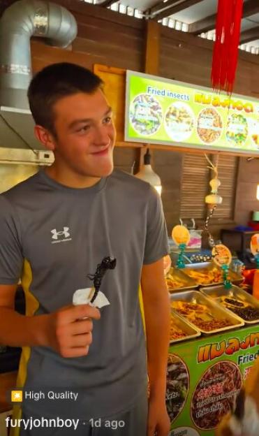 A young man holding a fried scorpion on a stick, with a sign for "Fried insects" in the background.