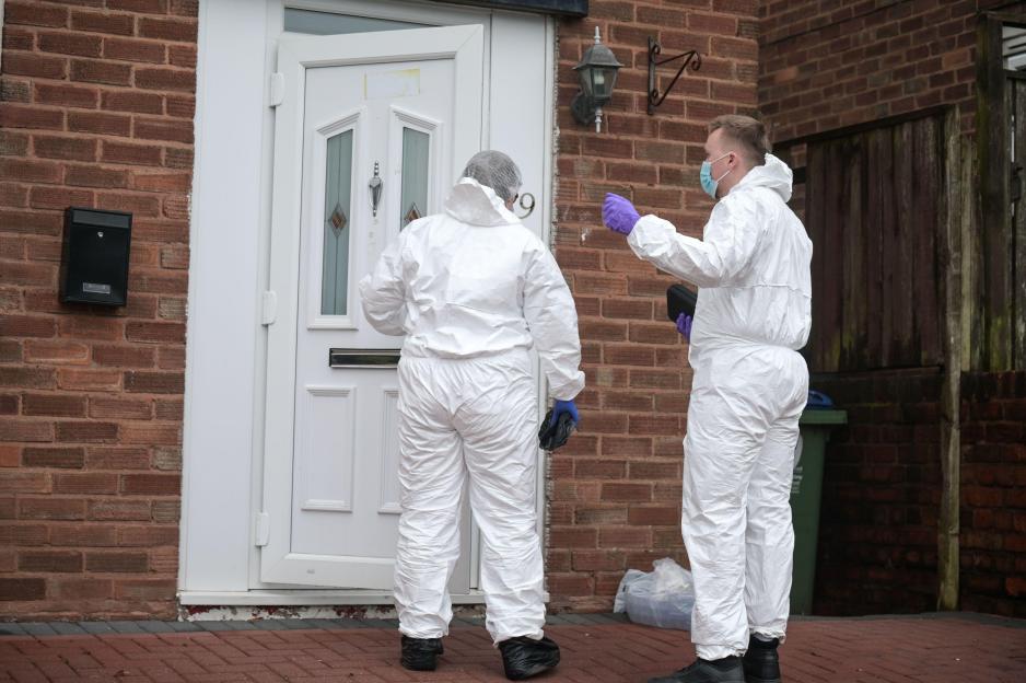 Two forensic officers in white suits stand outside the front door of a brick house.