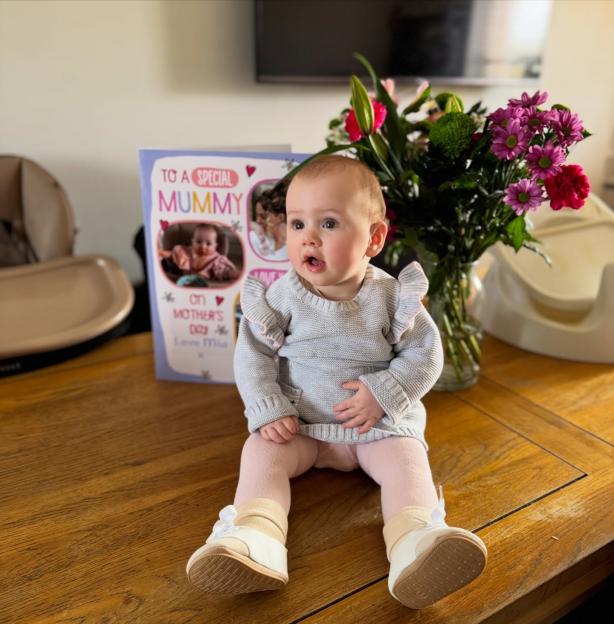 A baby sits on a wooden table next to a Mother's Day card and a bouquet of flowers.