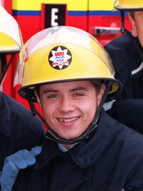 A young firefighter in a yellow helmet and dark uniform smiles at the camera.
